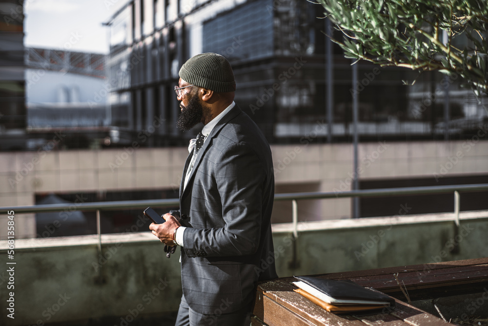 A profile shot of a middle-aged black man with an unshaven beard, eyeglasses, and a dark cap, holding a smartphone and leaning against a vase with an olive tree and a notebook sitting on top of it A profile shot of a middle-aged black man with an unshaven beard, eyeglasses, and a dark cap, holding a smartphone and leaning against a vase with an olive tree and a notebook sitting on top of it