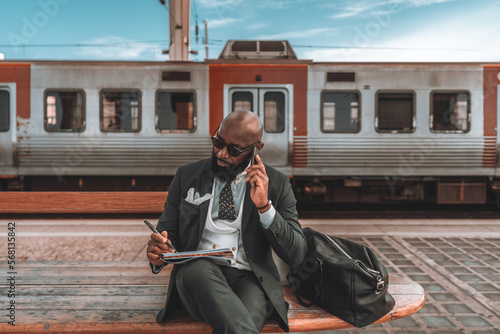 A black man multitasks on a blue-sky day, sitting on a wooden park bench while talking on the phone and jotting down notes Fototapet