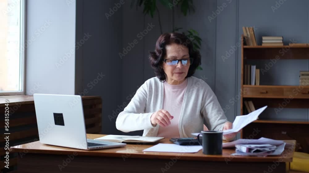 Middle aged senior woman sit with laptop and paper document. Pensive older mature lady reading paper bill pay online at home managing bank finances calculating taxes planning loan debt pension payment Middle aged senior woman sit with laptop and paper document. Pensive older mature lady reading paper bill pay online at home managing bank finances calculating taxes planning loan debt pension payment