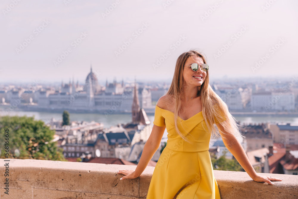 A young blonde tourist poses in front of the historic Parliament building on the Danube side in Budapest, Hungary, on a clear spring day