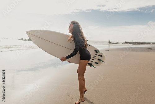 Full-lenght photo of exited lovely cute woman with loose wavy hair wearing black swimsuit having fun and enjoying vacation with surfboard on sandy beach with ocean view. 