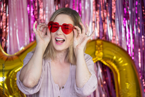 Woman with red love heart glasses smiling in front of pink streamer backdrop photo