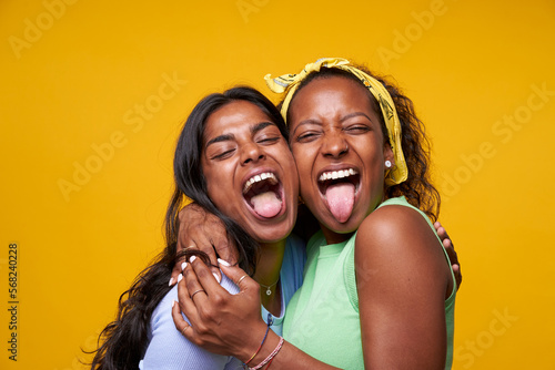 Two diversity pretty positive girls stick out your tongue hugging isolated on yellow color background. Young couple goofing around embracing and looking camera. Friendship and homosexual relationship. photo