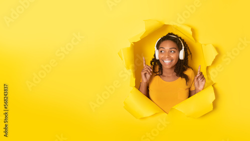 Joyful african american lady listening favourite music in wireless headphones, posing in paper hole on yellow background © Prostock-studio