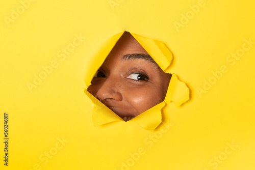 Young african american lady peeking out of a hole in yellow paper background, free copy space © Prostock-studio