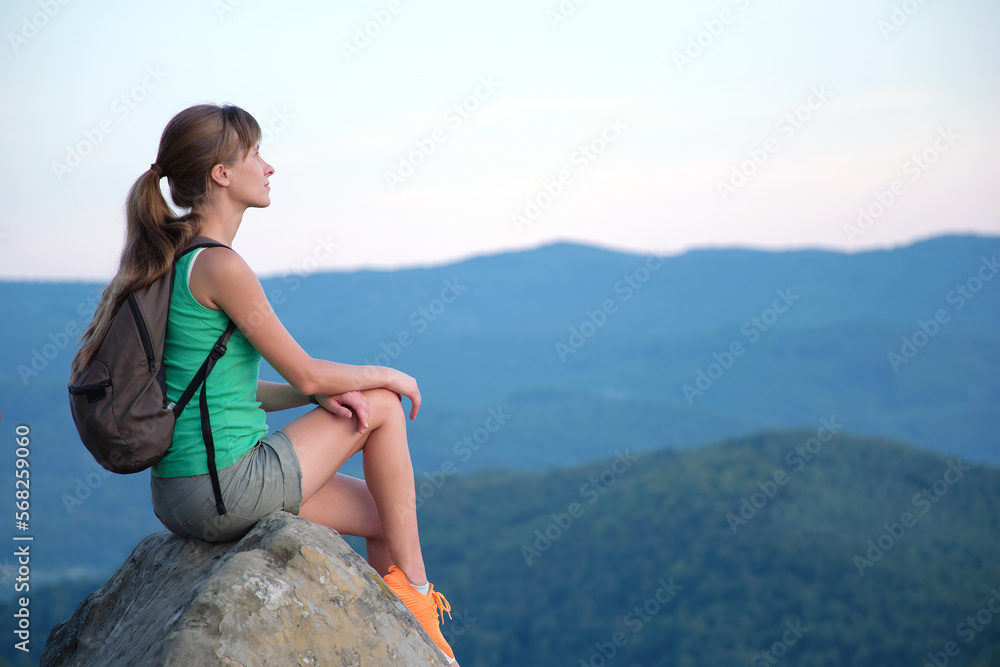 Naklejka premium Sportswoman sitting alone taking a break on hillside trail. Female hiker enjoying view of evening nature from rocky cliff on wilderness path. Active lifestyle concept