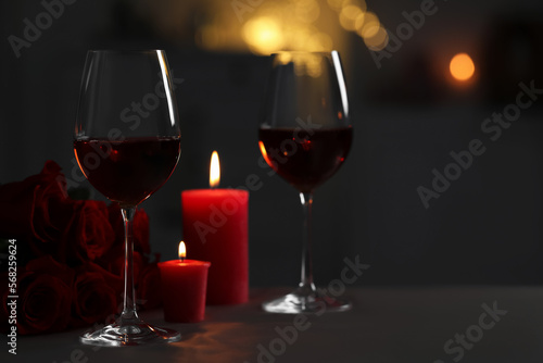Glasses of red wine, burning candles and rose flowers on grey table against blurred lights, space for text. Romantic atmosphere