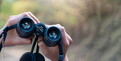 Closeup view of binoculars holding in hands of young asian natural surveyor, blurred dry autumn national forest park background.