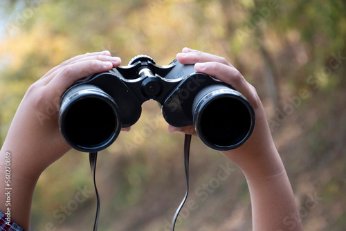 Closeup view of binoculars holding in hands of young asian natural surveyor, blurred dry autumn national forest park background.