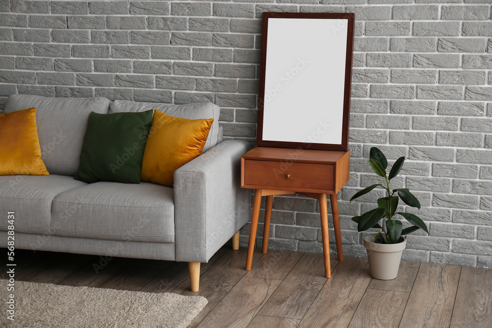 Naklejka premium Wooden table with blank frame, houseplant and sofa near grey brick wall
