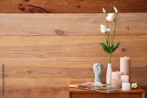 Burning candles, vase with eustoma flowers, magazine and decorative head on drawer near wooden wall