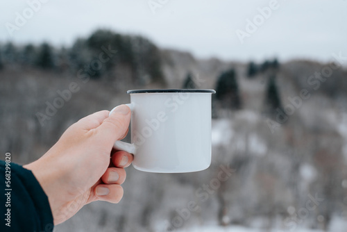 Close-up of caucasian male hand holding white enameled mug with an empty space for advertising on background of winter forest