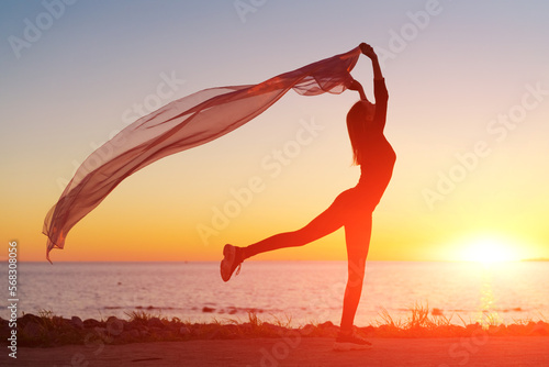 Silhouette slender girl at sunset on seashore rejoices with a transparent cloth in her hands in the wind. Emotional freedom concept