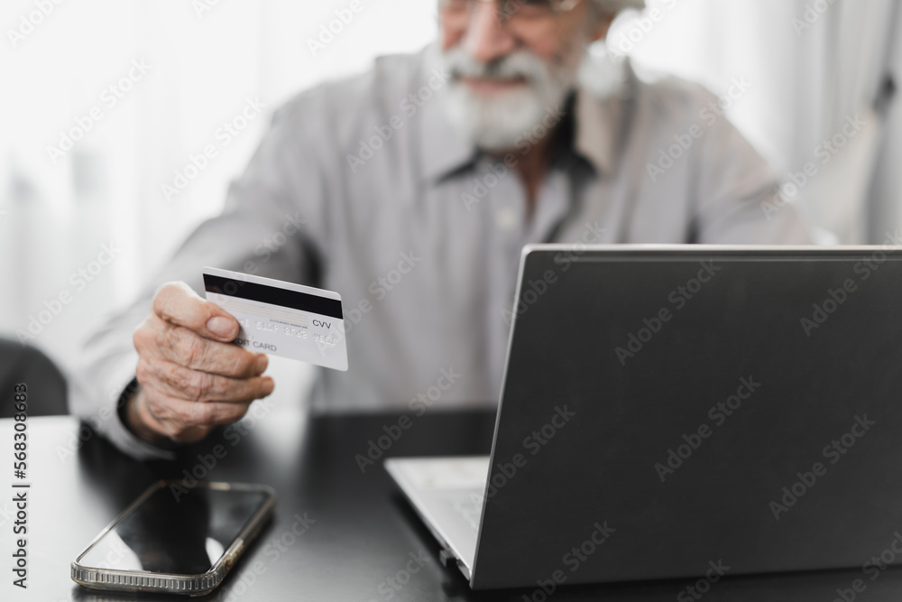 Caucasian Older man holding credit card and use computer notebook in leaving room at home Caucasian Older man holding credit card and use computer notebook in leaving room at home