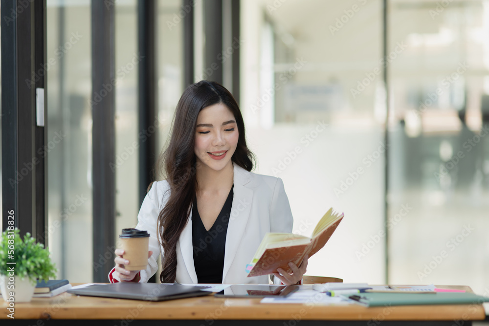 Friendly cheerful asian businesswoman reading a book and holding a coffee mug, people emotions and casual concept, positive woman working in office workspace. Friendly cheerful asian businesswoman reading a book and holding a coffee mug, people emotions and casual concept, positive woman working in office workspace.