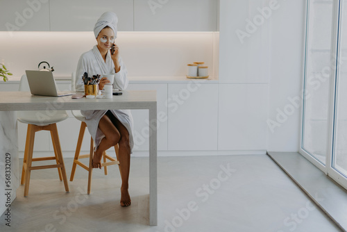 Pleased young woman dressed in bathrobe poses bare feet at chair in spacious kitchen sits at table with laptop and cosmetic products applies beauty patches under eyes talks via modern cellular © VK Studio