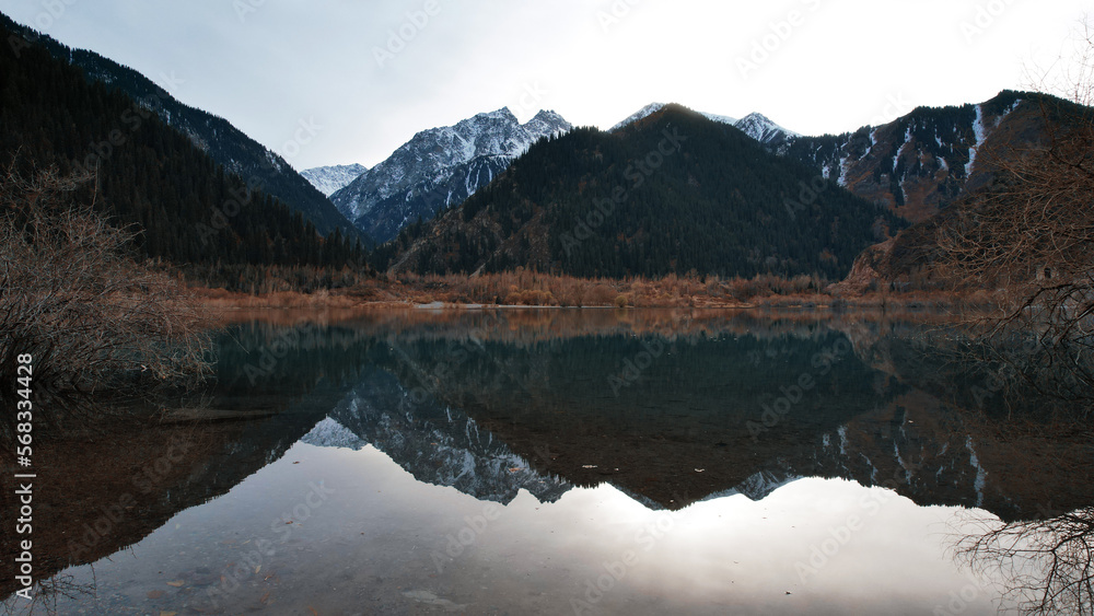 Issyk mountain lake with mirror water at sunset. The color of the water changes before our eyes. There are trees in clear water. Snowy mountains and green hills are visible. Clouds are reflected