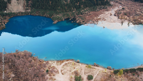 Turquoise blue mirror water with trees in the lake. Light streaks from underwater streams are visible. Autumn mountains and coniferous trees are reflected in the water. Issyk Mountain Lake, Kazakhstan
