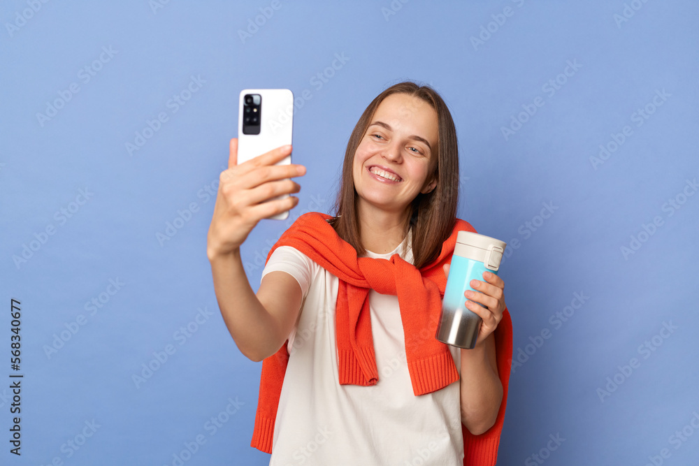 Image of smiling delighted Caucasian woman blogger wearing white T-shirt and orange sweater tied over shoulders standing isolated on blue background, taking selfie, making content for her blog. Image of smiling delighted Caucasian woman blogger wearing white T-shirt and orange sweater tied over shoulders standing isolated on blue background, taking selfie, making content for her blog.