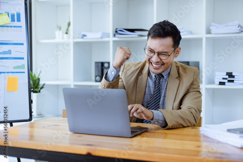 happy asian businessman and showing winner gesture while sitting at workplace in office, man very happy and excited doing winner. 	