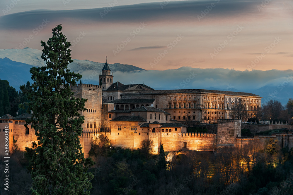 Palace of Carlos V in the Arab complex of the Alhambra in Granada, with Sierra Nevada and lenticular clouds in the background, illuminated with artificial light and a sunset sky.