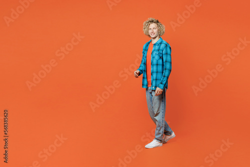 Full body sideways smiling fun young blond caucasian man wear blue shirt orange t-shirt look camera walking going strolling isolated on plain red background studio portrait. People lifestyle concept.