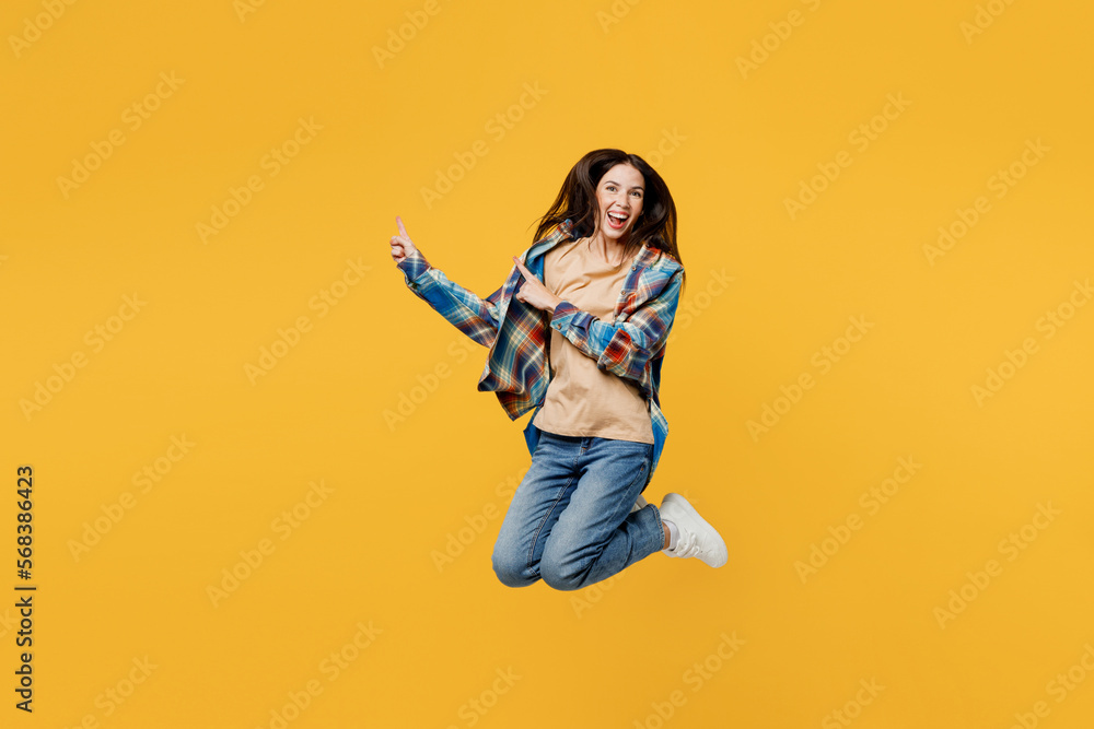Full body young smiling happy woman wear blue shirt beige t-shirt jump high point index finger aside indicate on workspace area copy space mock up isolated on plain yellow background studio portrait.