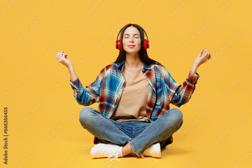 Full body young woman wearing blue shirt beige t-shirt headphones hold spreading hands in yoga om aum gesture relax meditate try to calm down listen music isolated on plain yellow background studio. Full body young woman wearing blue shirt beige t-shirt headphones hold spreading hands in yoga om aum gesture relax meditate try to calm down listen music isolated on plain yellow background studio.