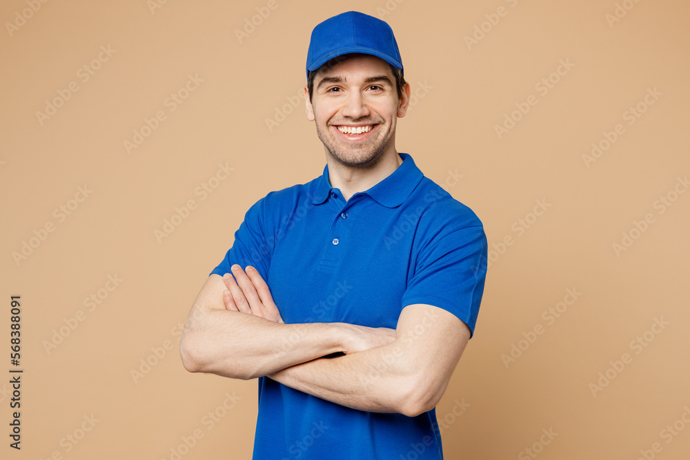 Professional delivery guy employee man wears blue cap t-shirt uniform workwear work as dealer courier look camera hold hands crossed folded isolated on plain light beige background. Service concept.