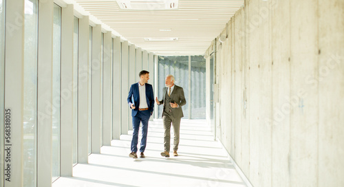 Young and a senior businessman walk down an office hallway, deep in conversation © BGStock72