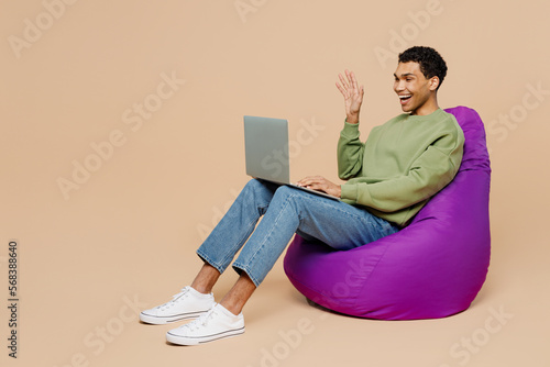 Full body young man of African American ethnicity wear green sweatshirt sit in bag chair work hold use laptop pc computer waving hand isolated on plain pastel light beige background studio portrait