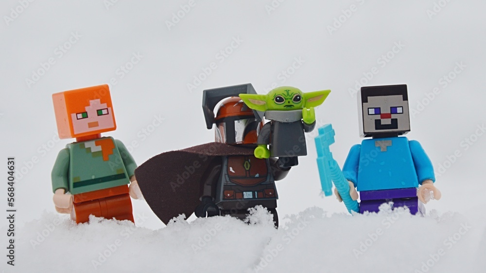 Fototapeta premium LEGO figures of Minecraft Steve and Alex standing together with LEGO Star Wars Mandalorian with baby Yoda, also known as Grogu on his left arm, in deep winter snow.