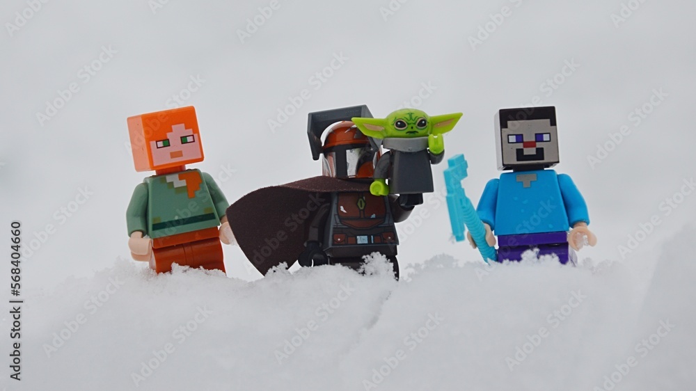 Naklejka premium LEGO figures of Minecraft Steve and Alex standing around LEGO Star Wars Mandalorian with baby Yoda, also known as Grogu on his left arm, in deep winter snow.