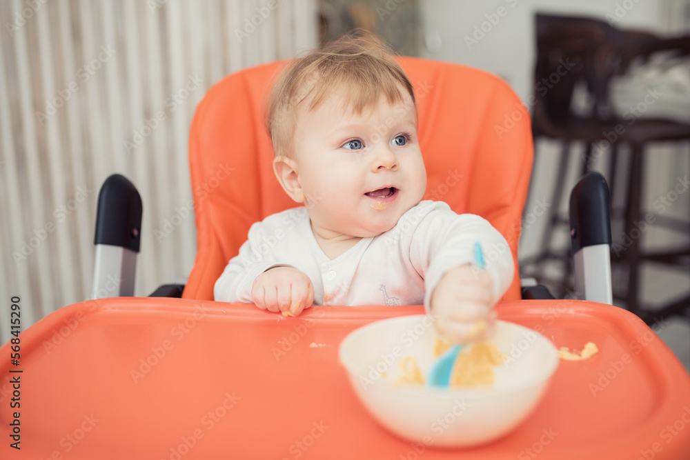 Fototapeta premium Cheerful happy child eats food by himself with a spoon. Portrait of a happy boy in a high chair for feeding.