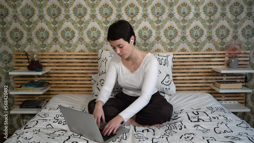 Beautiful woman is using her laptop while listening to music. The woman is sitting on the bed while she writes.