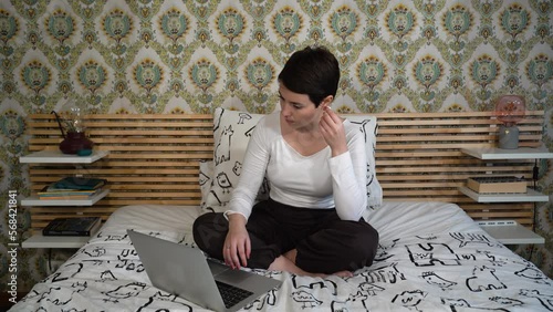 Beautiful woman is using her laptop while listening to music. The woman is sitting on the bed while she writes.