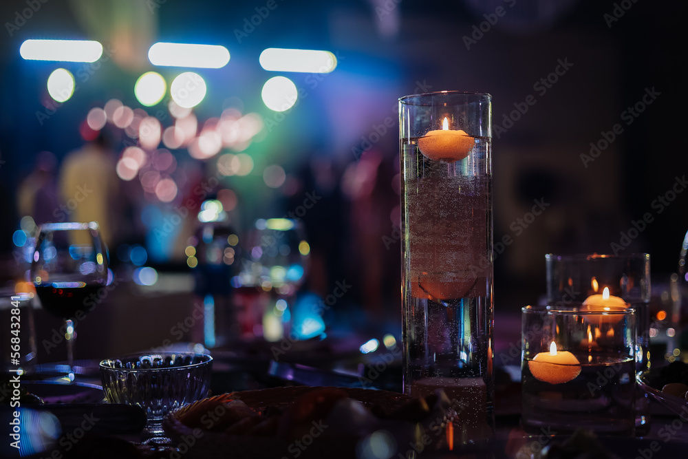 Banquet table decorated with burning candles in glass vases in restaurant hall. In the background party with silhouettes of people dancing on the dance floor with disco lights glowing searchlight