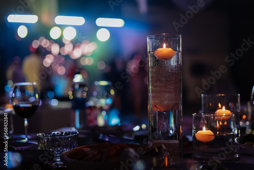 Banquet table decorated with burning candles in glass vases in restaurant hall. In the background party with silhouettes of people dancing on the dance floor with disco lights glowing searchlight