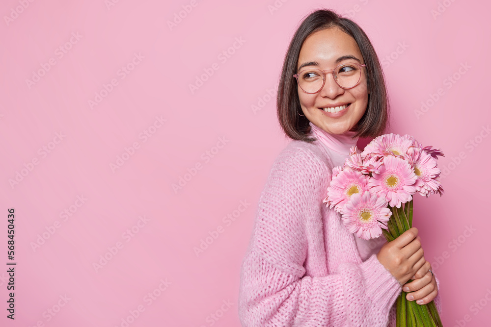 Studio shot of good looking woman smiles gently looks aside wears knitted loose jumper and spectacles holds boquet of gerberas likes receiving flowers isolated over pink background copy space for text