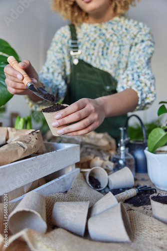 Cropped shot of unknown female gardener puts fetilized soil in pot for transplanting flower surrounded by different pots uses gardening tools stands near messy table. Horticulture and botaby concept