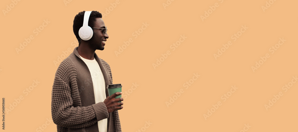 Portrait of stylish african man in wireless headphones listening to music with coffee cup looking away wearing knitted cardigan isolated on brown background, blank copy space for advertising text