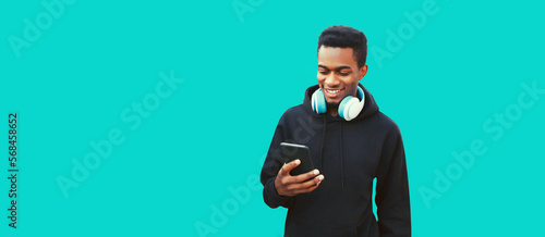 Portrait of happy smiling young african man with smartphone and headphones listening to music wearing black hoodie isolated on blue background, copy space