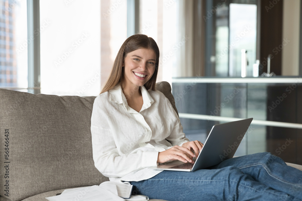 Happy beautiful young 20s laptop user woman typing on laptop, resting on big couch at in hotel room, looking at camera with toothy smile, working on project from home. Female portrait Happy beautiful young 20s laptop user woman typing on laptop, resting on big couch at in hotel room, looking at camera with toothy smile, working on project from home. Female portrait