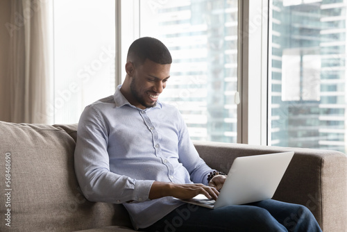 Happy young Black freelance professional man typing on laptop, sitting on couch at home, in modern hotel room with big window in background, smiling, laughing, having fun