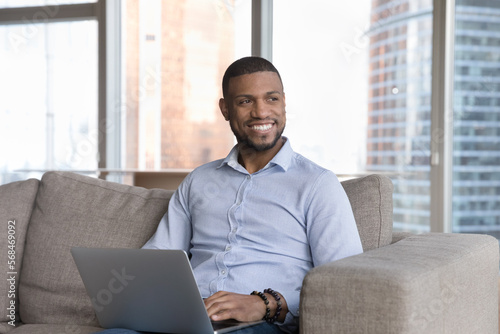 Happy attractive young businessman working on online freelance project from home, typing on laptop, sitting on couch, looking away, thinking over creative idea, smiling,