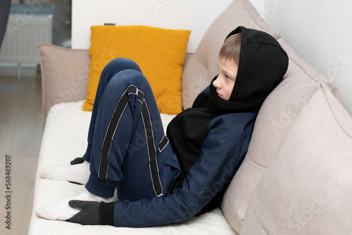People. Energy crisis concept. A teenage boy sits at home on the couch in gloves and a hood, freezes due to the heating being turned off. War in Europe. Energy crisis Ukraine.