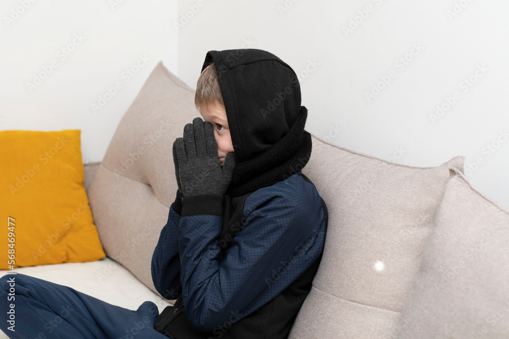 People. Energy crisis concept. A teenage boy sits at home on the couch in gloves and a hood, freezes due to the heating being turned off. War in Europe. Energy crisis Ukraine. People. Energy crisis concept. A teenage boy sits at home on the couch in gloves and a hood, freezes due to the heating being turned off. War in Europe. Energy crisis Ukraine.