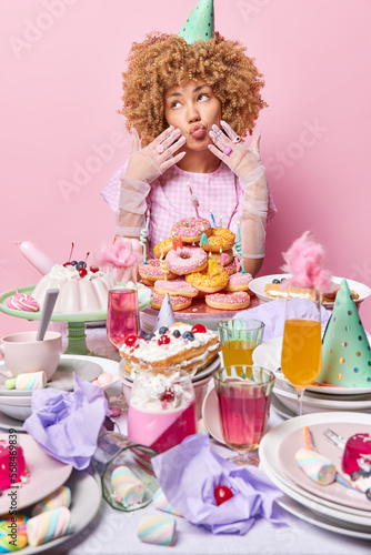 Dreamy curly haired woman wears cone hat dress and silk gloves keeps lips folded being deep in thoughts poses near table full of delicious desserts isolated over pink background. Celebration concept