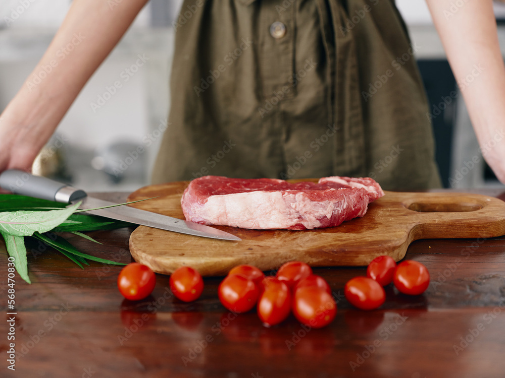 Woman with knife in hand cutting fresh steak meat for roasting in kitchen with salt pepper and other spices on table, red cherry tomatoes and herbs, preparing dinner. Woman with knife in hand cutting fresh steak meat for roasting in kitchen with salt pepper and other spices on table, red cherry tomatoes and herbs, preparing dinner.