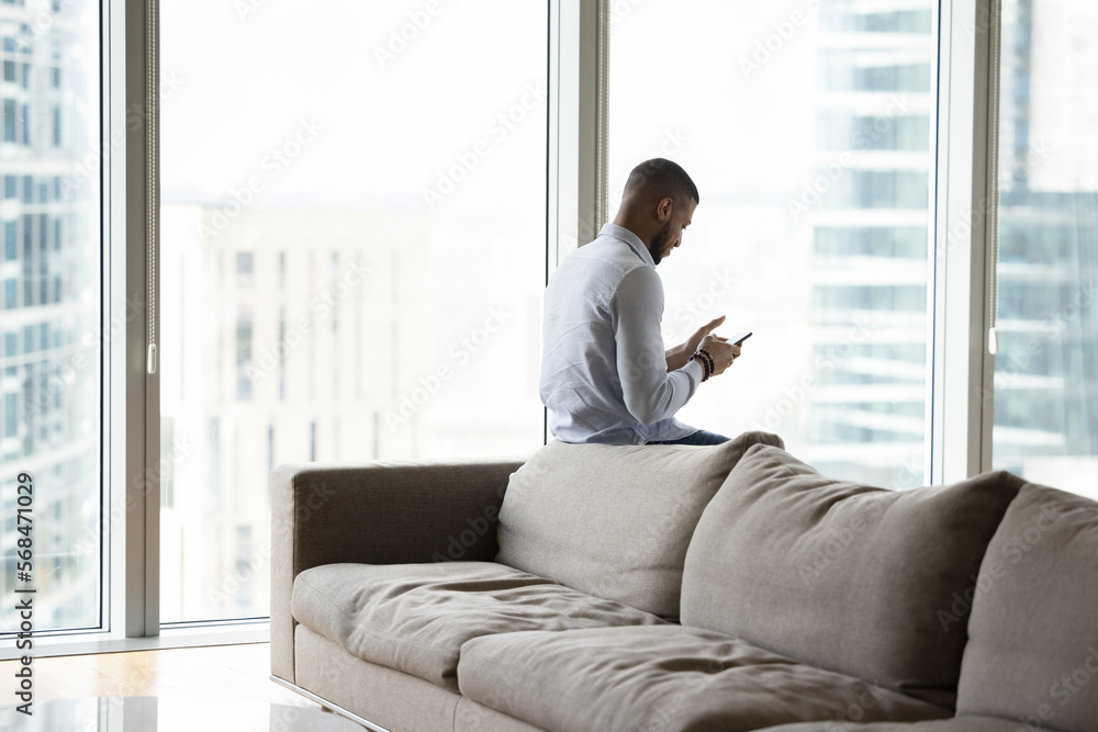 Focused young Black cellphone user man using digital gadget for Internet communication at home, chatting over work business project, typing on mobile phone, leaning on soft couch at big urban window Focused young Black cellphone user man using digital gadget for Internet communication at home, chatting over work business project, typing on mobile phone, leaning on soft couch at big urban window
